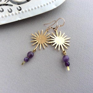 May include: A pair of gold-colored earrings featuring a sunburst design. Each earring has a sun-shaped pendant with multiple rays and is adorned with two purple amethyst beads. The earrings hang from gold-colored ear wires.