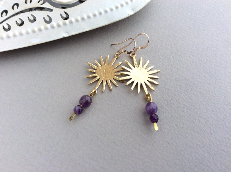 May include: A pair of gold-colored earrings featuring a sunburst design. Each earring has a sun-shaped pendant with multiple rays and is adorned with two purple amethyst beads. The earrings hang from gold-colored ear wires.