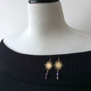 May include: Gold-colored sunburst earrings with dangling purple beads. The earrings are displayed on a black ribbed top, showcasing their design and color contrast. The sunburst design has multiple rays extending outwards.