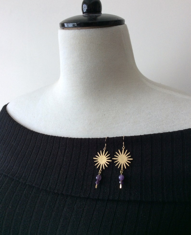 May include: Gold-colored sunburst earrings with dangling purple beads. The earrings are displayed on a black ribbed top, showcasing their design and color contrast. The sunburst design has multiple rays extending outwards.