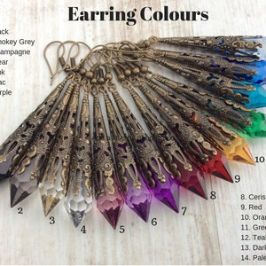 May include: A collection of long, ornate earrings in various colors, including black, smokey grey, and purple. Each earring features a faceted crystal pendant encased in a detailed, antique bronze filigree setting. The image includes the text "Earring Colours".