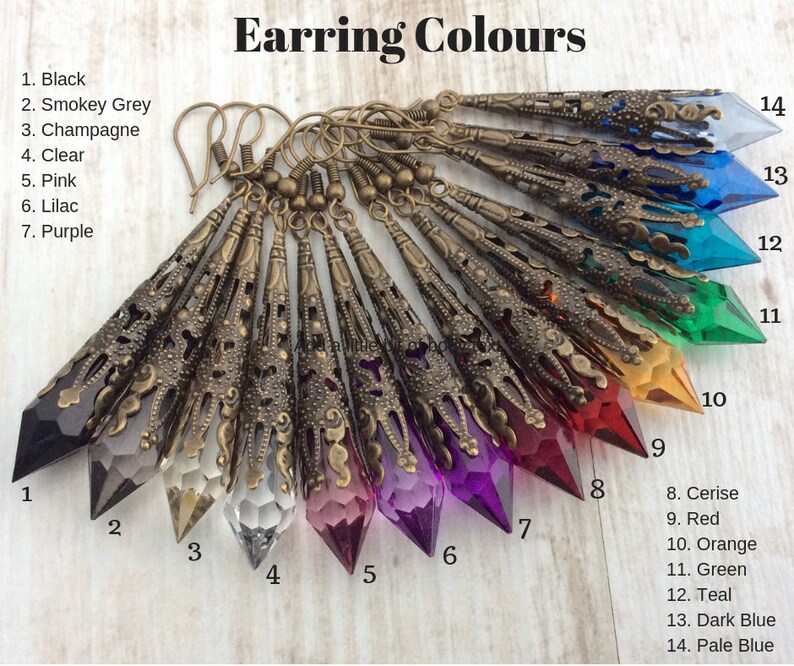 May include: A collection of long, ornate earrings in various colors, including black, smokey grey, and purple. Each earring features a faceted crystal pendant encased in a detailed, antique bronze filigree setting. The image includes the text "Earring Colours".