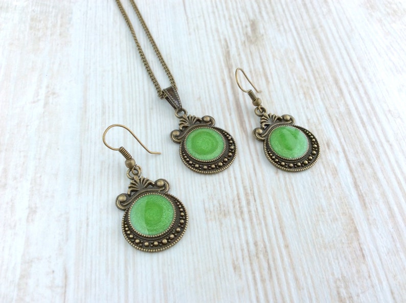 May include: A jewelry set featuring a necklace and earrings. The set has a bronze-colored metal with a decorative design. Each piece has a round, bright green center. The earrings have a hook closure.