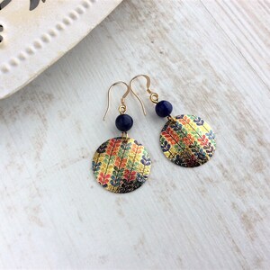 May include: Gold-tone earrings with a circular, patterned design. Each earring features a navy blue bead and a round, gold-colored disc adorned with colorful leaf patterns in red, yellow, and green. The earrings are displayed on a light-colored wooden surface.