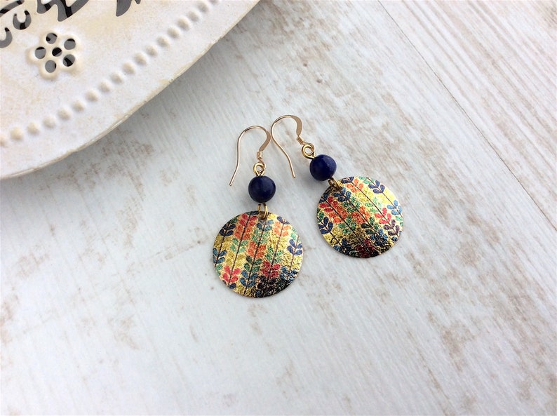 May include: Gold-tone earrings with a circular, patterned design. Each earring features a navy blue bead and a round, gold-colored disc adorned with colorful leaf patterns in red, yellow, and green. The earrings are displayed on a light-colored wooden surface.