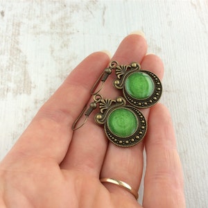 May include: A pair of antique-style earrings with a bronze finish. Each earring features a round, vibrant green center surrounded by a detailed, ornate bronze frame. The earrings are held in a hand, showcasing their size and design.