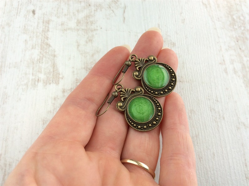 May include: A pair of antique-style earrings with a bronze finish. Each earring features a round, vibrant green center surrounded by a detailed, ornate bronze frame. The earrings are held in a hand, showcasing their size and design.