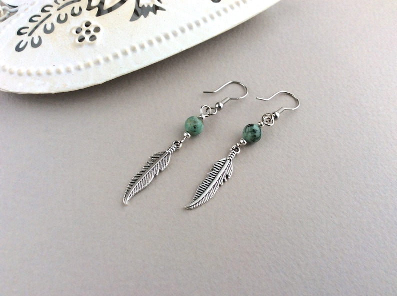 May include: A pair of dangle earrings featuring silver-toned feather charms and small, round, green beads. The earrings have a hook closure and are displayed on a light gray surface.