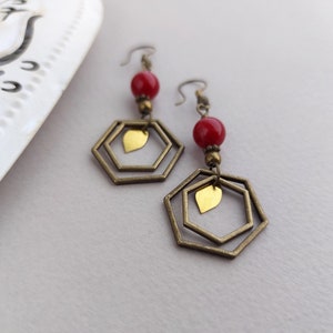 May include: A pair of dangle earrings featuring red beads, brass-colored hexagonal frames, and small gold-colored leaf charms. The earrings have a vintage aesthetic and are displayed against a neutral background.