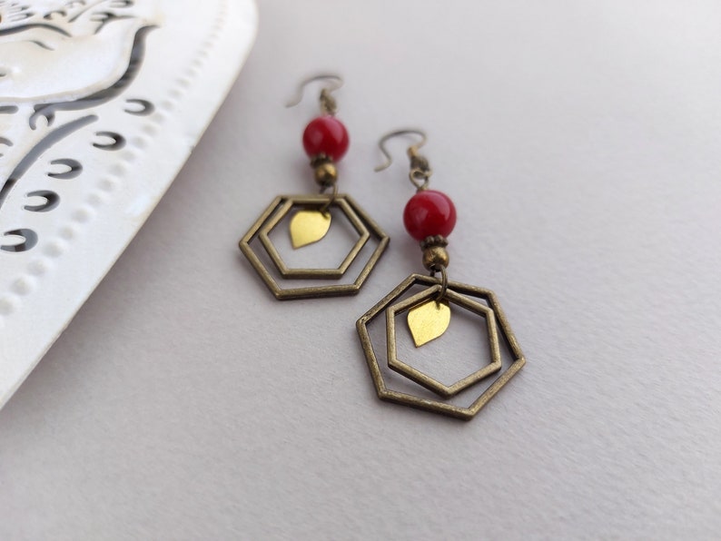 May include: A pair of dangle earrings featuring red beads, brass-colored hexagonal frames, and small gold-colored leaf charms. The earrings have a vintage aesthetic and are displayed against a neutral background.