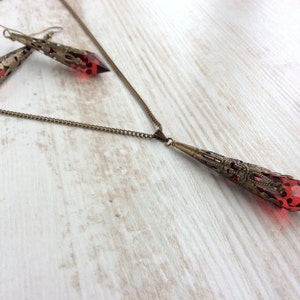 May include: A vintage-style jewelry set featuring a necklace and earrings. The necklace has a long, ornate pendant with a faceted red gemstone. The earrings mirror the pendant design. All pieces have a bronze-colored metal finish.