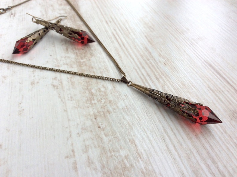 May include: A vintage-style jewelry set featuring a necklace and earrings. The necklace has a long, ornate pendant with a faceted red gemstone. The earrings mirror the pendant design. All pieces have a bronze-colored metal finish.