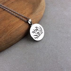 Puede incluir: Un collar plateado con un colgante circular. El colgante presenta un dibujo lineal negro de plantas estilizadas. La cadena es delicada y descansa sobre una superficie de madera.