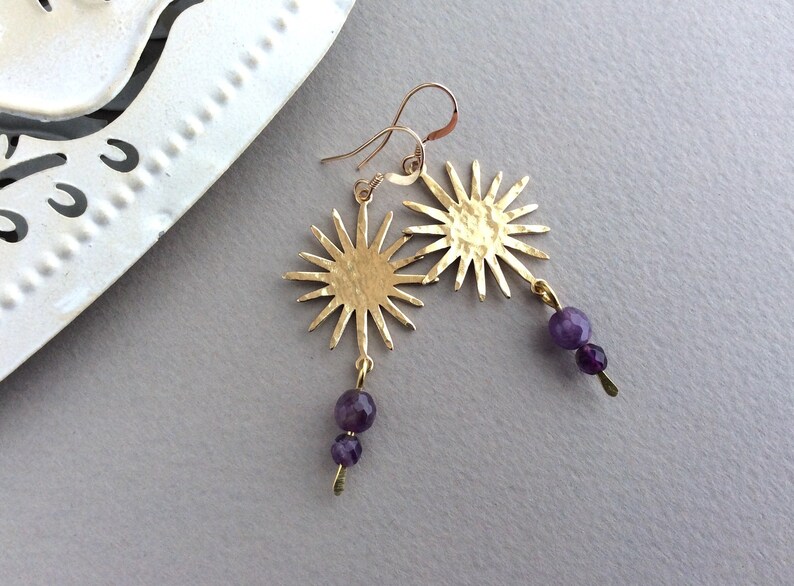 May include: Gold-tone sunburst earrings with a hammered texture. Each earring features a sun design and dangles with two faceted purple beads. The earrings are displayed on a light gray surface.