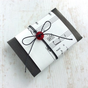 May include: A small, rectangular gift wrapped in black paper and sheet music. The package is tied with black string and a red button. The sheet music has musical notes and the word "want" visible.