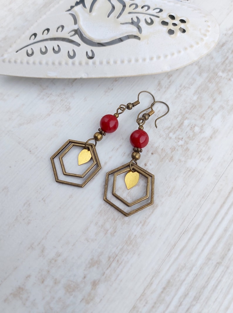 May include: A pair of dangle earrings featuring red beads, brass-colored hexagonal frames, and small gold-colored leaf charms. The earrings have a vintage aesthetic and are designed for pierced ears.
