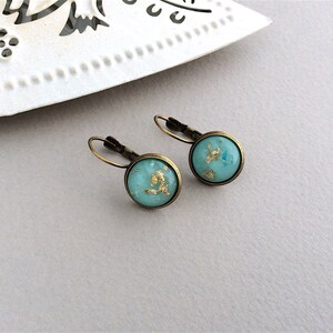May include: A pair of round earrings with a turquoise blue background and gold leaf accents. The earrings have a bronze-colored metal frame and lever-back closures. The earrings are displayed on a light gray surface.
