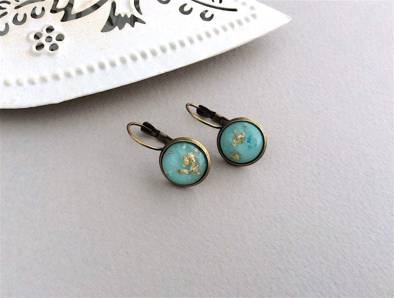 May include: A pair of round earrings with a turquoise blue background and gold leaf accents. The earrings have a bronze-colored metal frame and lever-back closures. The earrings are displayed on a light gray surface.