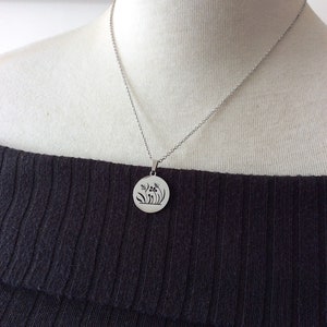 Puede incluir: Un collar con colgante plateado con un dije circular con un dise&ntilde;o floral. El collar se muestra en un maniqu&iacute; que lleva un top negro con los hombros descubiertos. El colgante presenta flores y hojas estilizadas.