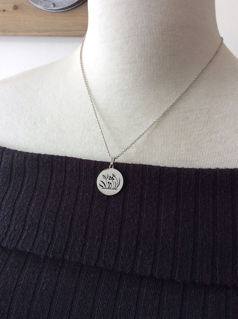 Puede incluir: Un collar con colgante plateado con un dije circular con un dise&ntilde;o floral. El collar se muestra en un maniqu&iacute; que lleva un top negro con los hombros descubiertos. El colgante presenta flores y hojas estilizadas.