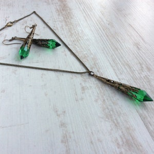 May include: A necklace and earrings set featuring green crystal pendants with bronze-colored filigree accents. The necklace has a long chain with a matching pendant. The earrings have a similar design, with a hook closure.