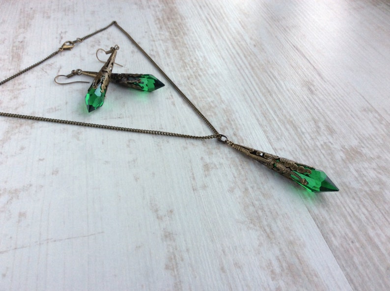 May include: A necklace and earrings set featuring green crystal pendants with bronze-colored filigree accents. The necklace has a long chain with a matching pendant. The earrings have a similar design, with a hook closure.
