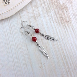 May include: A pair of dangle earrings featuring a red bead and a silver-colored feather charm. The earrings have a hook closure and are displayed on a light wooden surface. The jewelry is simple and elegant.