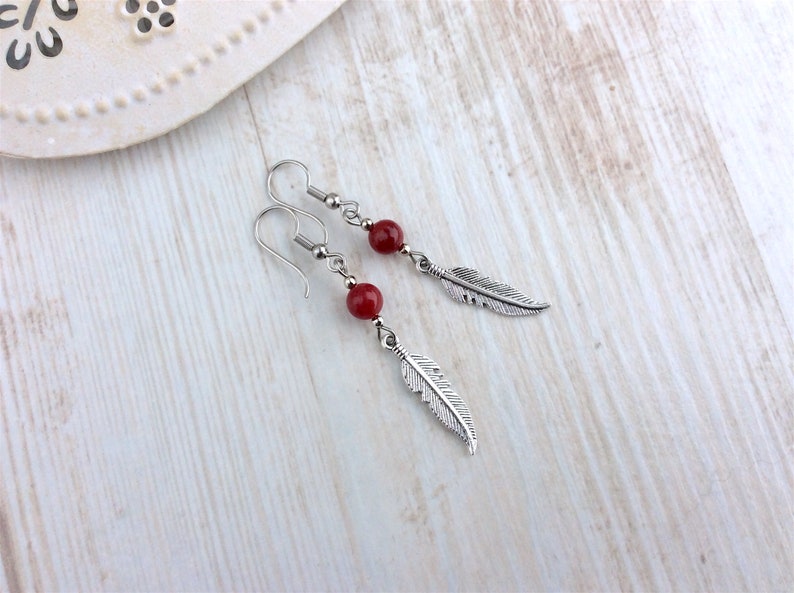 May include: A pair of dangle earrings featuring a red bead and a silver-colored feather charm. The earrings have a hook closure and are displayed on a light wooden surface. The jewelry is simple and elegant.