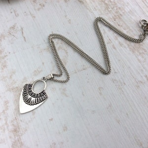 May include: A silver-toned necklace with a pendant featuring an oval and a stylized, decorative bottom edge. The chain is a fine, linked design. The necklace is displayed on a light-colored, wood-grain surface.