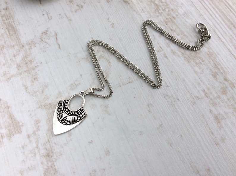 May include: A silver-toned necklace with a pendant featuring an oval and a stylized, decorative bottom edge. The chain is a fine, linked design. The necklace is displayed on a light-colored, wood-grain surface.