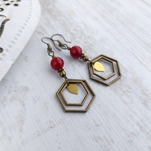 May include: A pair of dangle earrings featuring red beads, brass-colored metal accents, and geometric hexagon shapes. The earrings have a leaf-shaped charm within the hexagon design, and are on a white wooden surface.