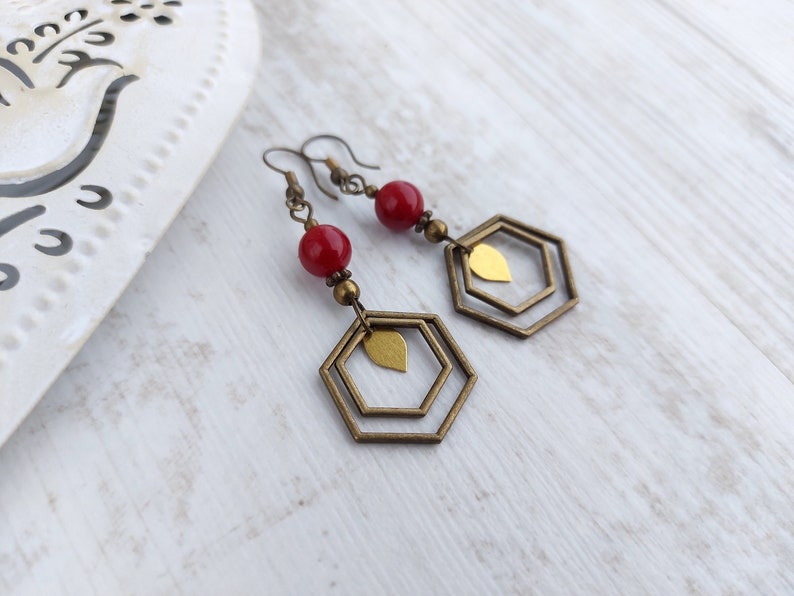 May include: A pair of dangle earrings featuring red beads, brass-colored metal accents, and geometric hexagon shapes. The earrings have a leaf-shaped charm within the hexagon design, and are on a white wooden surface.