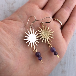 May include: Gold-tone sunburst earrings with purple amethyst beads. The earrings feature a sun design with multiple rays and a small, dangling cluster of purple beads. The earrings are displayed against a neutral background.
