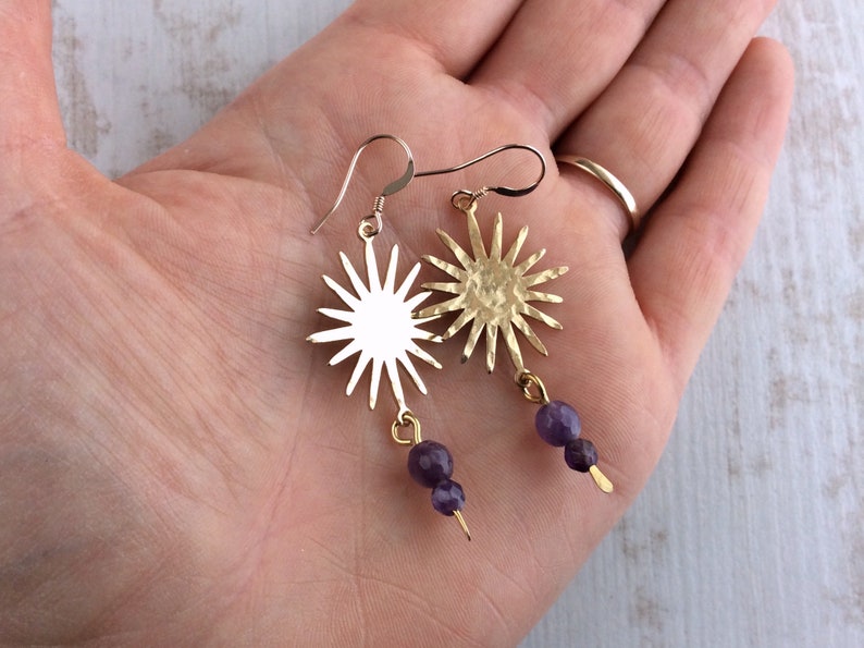 May include: Gold-tone sunburst earrings with purple amethyst beads. The earrings feature a sun design with multiple rays and a small, dangling cluster of purple beads. The earrings are displayed against a neutral background.