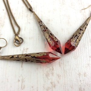 May include: A jewelry set featuring a necklace and earrings with red crystal pendants. The pendants are encased in ornate, antique bronze filigree. The necklace has a matching chain and clasp, and the earrings have hook closures.