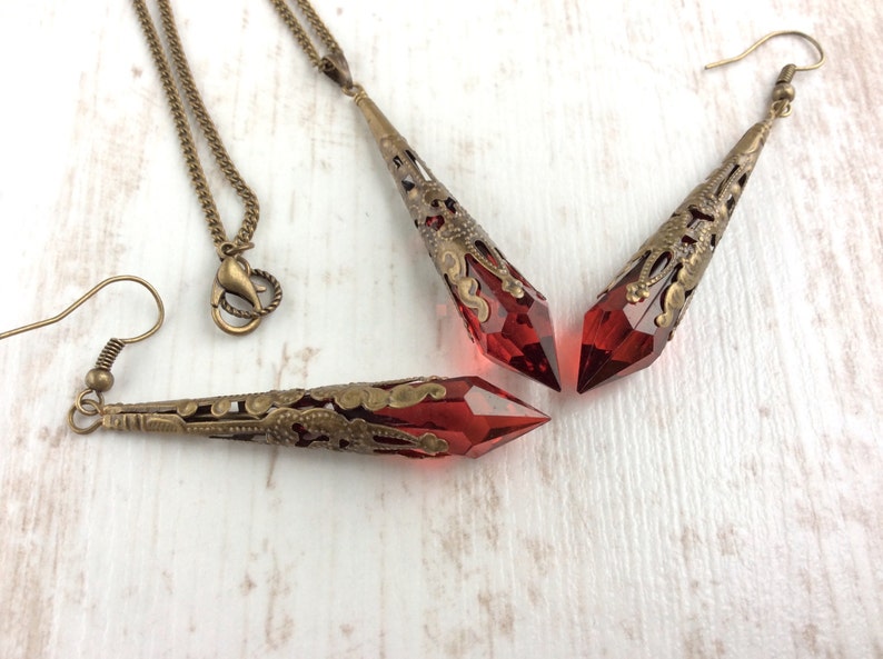 May include: A jewelry set featuring a necklace and earrings with red crystal pendants. The pendants are encased in ornate, antique bronze filigree. The necklace has a matching chain and clasp, and the earrings have hook closures.