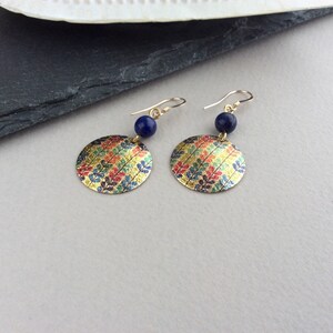 May include: A pair of gold-colored earrings with a circular, patterned design. Each earring features a gold hook, a small, dark blue bead, and a flat, round pendant with a colorful leaf pattern in gold, red, blue, and green.