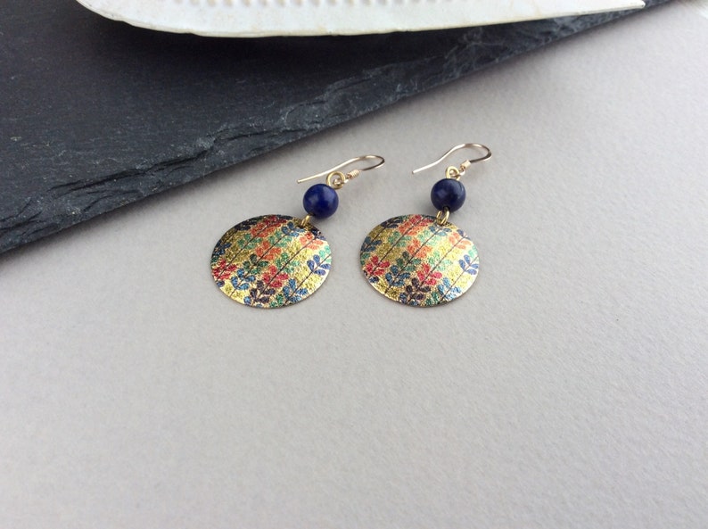 May include: A pair of gold-colored earrings with a circular, patterned design. Each earring features a gold hook, a small, dark blue bead, and a flat, round pendant with a colorful leaf pattern in gold, red, blue, and green.