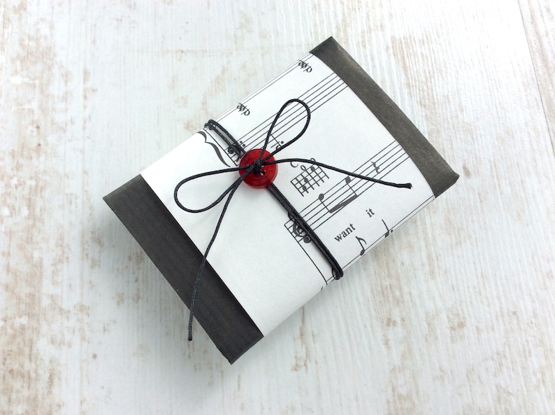 May include: A small, wrapped package with a sheet music design. The package is wrapped in black paper and tied with black string, secured with a red button. The sheet music has the words "want it" printed on it.