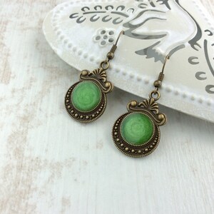 May include: A pair of antique-style earrings with a green center and bronze-colored filigree detailing. The earrings have a circular design with a textured green center and a decorative border. The ear hooks are also bronze-colored.
