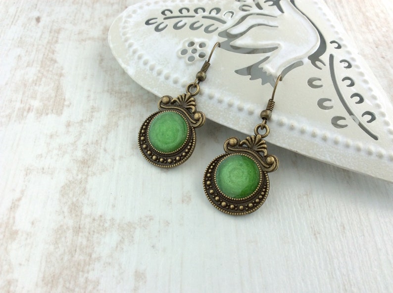 May include: A pair of antique-style earrings with a green center and bronze-colored filigree detailing. The earrings have a circular design with a textured green center and a decorative border. The ear hooks are also bronze-colored.