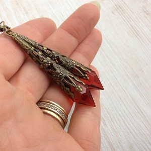May include: Long, antique brass-toned earrings with a filigree design and faceted, red crystal pendants. The earrings have a vintage aesthetic and are suitable for special occasions or everyday wear. The earrings are approximately 3 inches long.