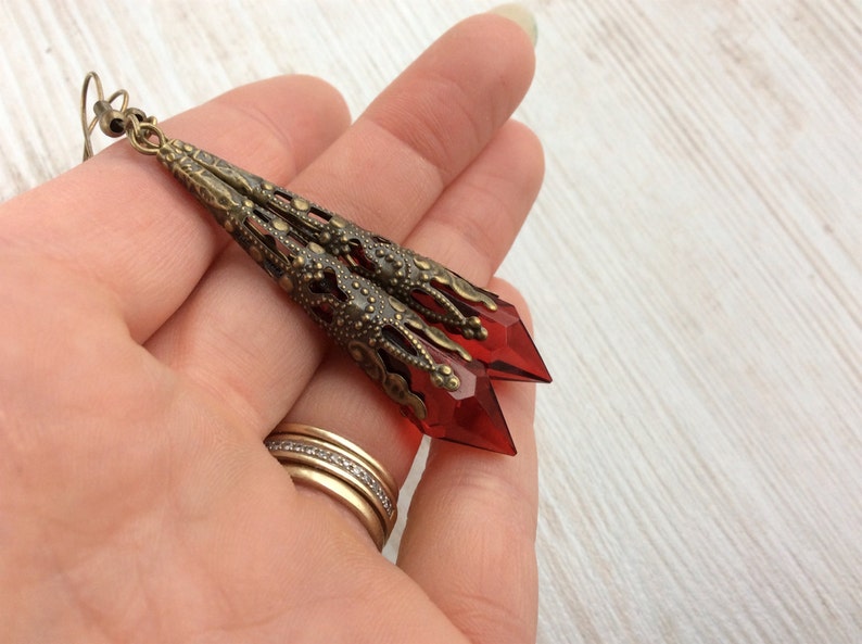 May include: Long, antique brass-toned earrings with a filigree design and faceted, red crystal pendants. The earrings have a vintage aesthetic and are suitable for special occasions or everyday wear. The earrings are approximately 3 inches long.