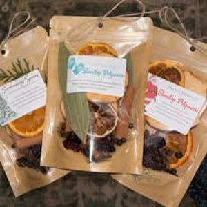 May include: Three stand-up pouches made of brown kraft paper with clear windows, each containing dried orange slices, cinnamon sticks, bay leaves, and dark berries. Each pouch features a white label with text and a string tie at the top.