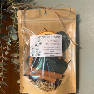 May include: A clear pouch with a brown paper backing containing stovetop potpourri. The label reads Breathe Easy Stovetop Potpourri, with instructions to pour contents into a small pot, cover with water, and simmer. Visible ingredients include dried orange slices, cinnamon sticks, dark leaves, and herbs.