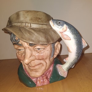 May include: A ceramic pitcher shaped like a man's head wearing a brown hat with a fish sticking out of the top. The man has a green jacket and a red shirt.
