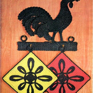 Pode incluir: Um galo preto de crochê pendurado na parede com dois pegadores de panela de crochê, um amarelo e um vermelho, pendurados abaixo.