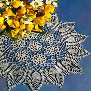 Puede incluir: Un encaje de ganchillo blanco con un borde festoneado y un diseño floral en el centro. El encaje está sobre un fondo azul y un ramo de flores amarillas y blancas está en la esquina superior izquierda.
