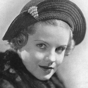 May include: A black and white portrait of a young woman wearing a black hat with a decorative pin and a fur collar.