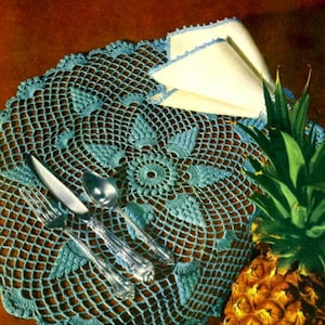 Puede incluir: Un encaje de ganchillo azul turquesa con un delicado patrón floral. El encaje está colocado plano sobre una superficie marrón y presenta un juego de cubiertos y una piña.